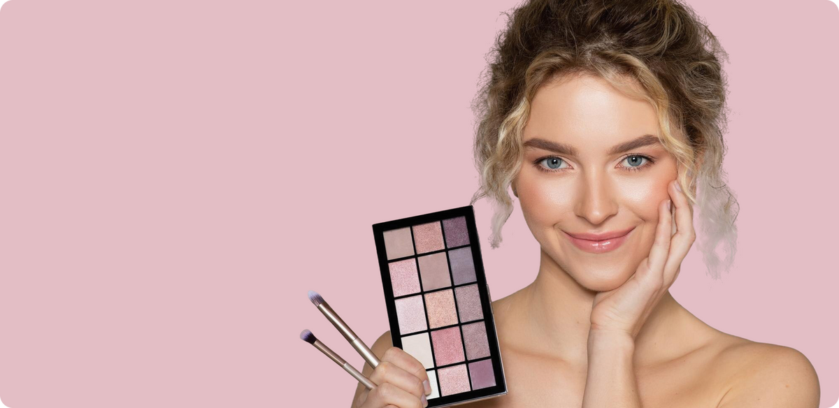 A woman with styled blonde hair posing on a solid pink background, holding a 12-shade eyeshadow palette and two makeup brushes.